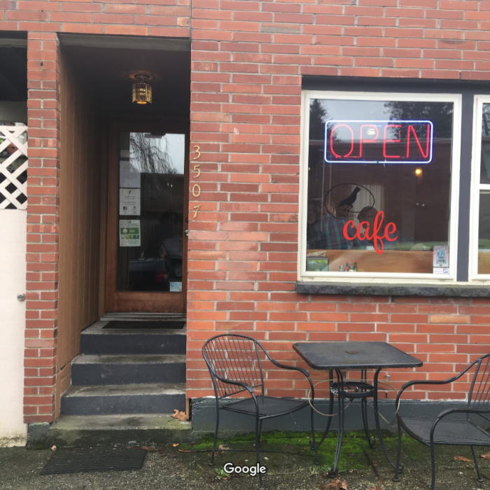 The entrance to a cafe with a table outside, surrounded in brick with a neon welcome sign in the window.